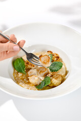 Woman hand holding fork with sea scallop over creamy risotto. Person eat seafood risotto with scallops. Female hand with italian risotto. Trendy menu concept.