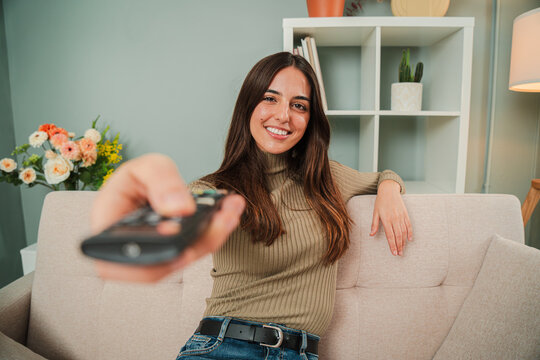 One young woman using the television remote controller to change the channel. Adult caucasian female turning on the smart tv to watch a movie or show. Smiley girl pressing the volume control button