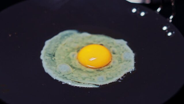 A chef fries an egg, sunny side up, in a small frying pan over an industrial gas burning stove
