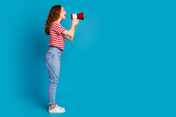 Full size photo of adorable girl wear stylish t-shirt denim trousers scream in megaphone to empty space isolated on blue color background