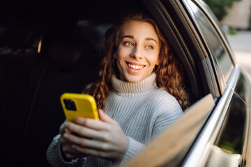 Happy woman using smartphone, sitting in the back seat of a car, scrolling social media news,...