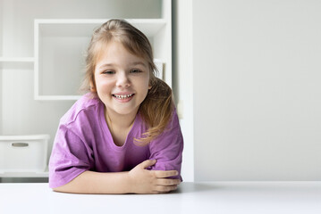 Caucasian child girl portrait sitting at white table empty space background. Product display design.Little kid girl happy smiling face.