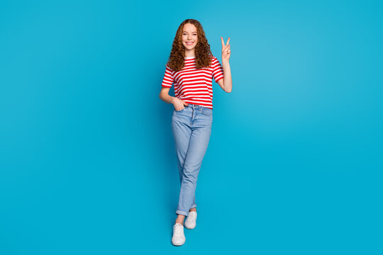 Full size photo of adorable girl wear stylish t-shirt denim trousers showing v-sign symbol arm in pocket isolated on blue color background