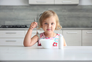 Little caucasian girl sitting at kitchen table with spoon and bowl, child has a meal, breackfast. Kid's nutrition concept.
