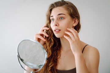 Young woman with red spot unhappy about acne and skin imperfections, looking at reflection in mirror in bathroom. Facial skin care. Acne problem.