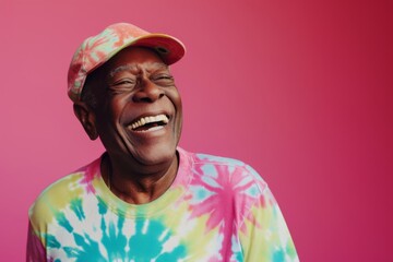 A cheerful African American senior man wearing a tie-dye shirt and cap, radiating positivity against a vibrant pink background.