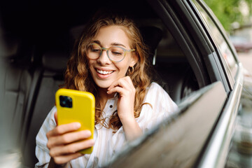 Young woman uses a smartphone while sitting in the back seat of a car. Female happy in car...