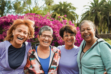 Group of happy diverse senior women hugging each other at city park while smiling on camera -...
