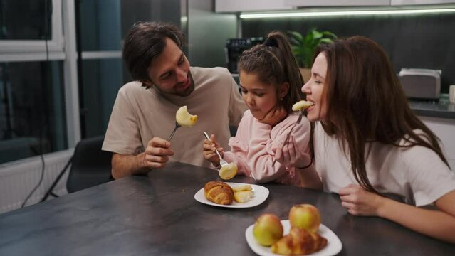 A happy brunette man with stubble in a beige T-shirt together with his brunette wife in a white T-shirt and a little daughter in a pink suit eat cheesecakes during their breakfast in a modern