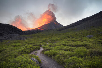 Smoke billows from a volcanic peak, casting a red glow across the lush green mountain valleys below