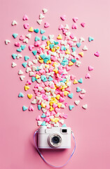 A white mini camera with lots of colorful candy hearts spilling out the top, on a pastel pink background, in a minimalistic style