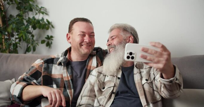 Happy gay couple a middle-aged man with gray hair and a lush beard takes a selfie using a White phone along with his brunette boyfriend in a plaid shirt while sitting on the sofa in a modern apartment