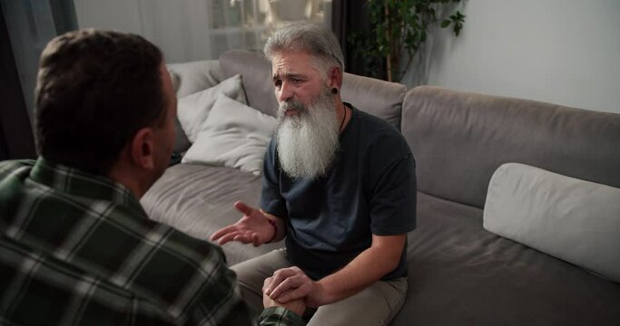Over the shoulder A sad man with gray hair and a lush gray beard in a gray T-shirt communicates with his boyfriend a brunette with stubble in a checkered shirt and holds his hands while sitting on a