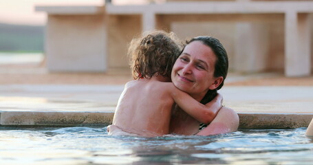 Small boy hugging mother inside swimming pool water. Toddler child embracing mom