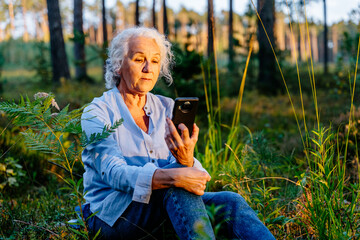 Elderly healthy life concept. An advanced modern grandmother sits in meadow in the woods talking on...