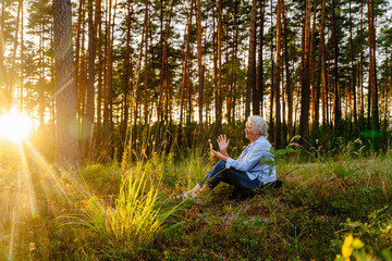 Elderly healthy life concept. Beautiful senior woman sit at forest meadow at sunset, waving hand, having video call, admiring nature with pine woods on background.