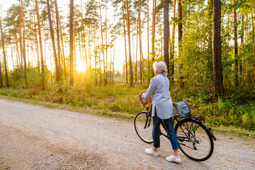 Active life in golden years concept. Senior woman pushes bike along forest path at summer sunset....