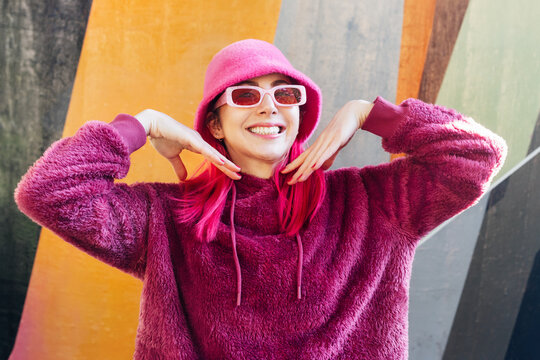Smiling young woman with pink hair and sunglasses in a magenta fluffy sweatshirt and bucket hat on the wall background. Urban hipster street fashion. Mono color look. Vanilla Girl. Kawaii vibes.
