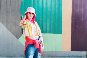 Young woman with pink hair and sunglasses in Bucket hat and multicolor strippled shirt making V...