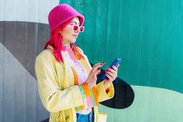 Stylish young woman using her phone for messaging, communication or content creating for social media standing on graffiti wall background. Bright hipster street fashion. Influencer, blogger. - obrazy, fototapety, plakaty