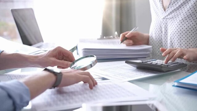 Business colleagues, accountants, or auditors are reviewing financial data in fair office. Two professionals are analyzing paper reports and using calculators, focusing on aspects of audit and taxes