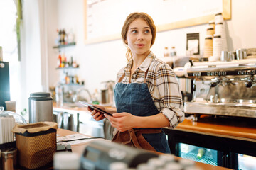 A young woman in an apron with a digital tablet at the bar counter of a coffee shop takes an order....