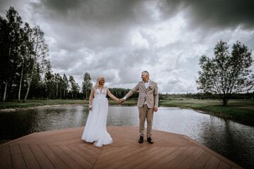 Valmiera, Latvia - Augist 13, 2023 - A bride and groom hold hands on a wooden pier extending into a...