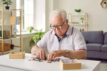 Portrait of a senior elderly sad gray-haired man collecting wooden jigsaw puzzles game at home...