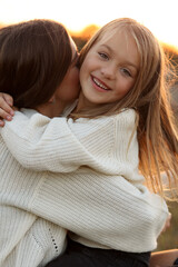 Family day, mother's day. Happy smiling young mom and adorable child daughter soft hugging, kissing and spending time together at autumn on sunset. Idyllic family having fun outdoors on fall holiday.