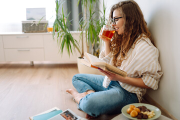 Young woman at home drinking coffee or tea and reading book or magazine. Lifestyle, relaxation and domestic life, comfort concept.
