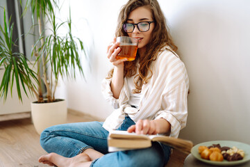 Young woman at home drinking coffee or tea and reading book or magazine. Lifestyle, relaxation and domestic life, comfort concept.