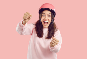 Crazy smiling amazed woman with opened mouth in helmet imagines she is driving and holding steering wheel on pink background. She is wearing pink sweatshirt. Human emotions, energy, drive concept. - obrazy, fototapety, plakaty