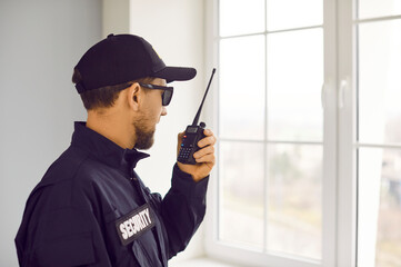 Security guard man in uniform cap, jacket and sunglasses is standing by window inside house and talking on portable wireless two way walkie talkie transceiver radio set device. Property safety concept - 776258783
