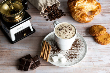 Hot coffee mug with foam, coffee beans and croissant in background