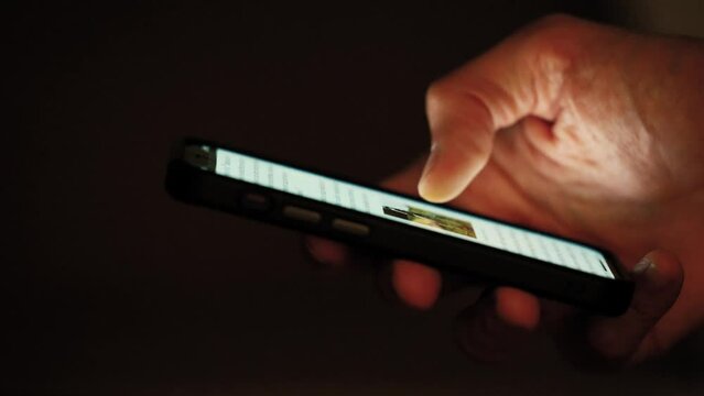 Close-Up Hand Scrolling Smartphone at Night, Dark Background Lit by Screen Light