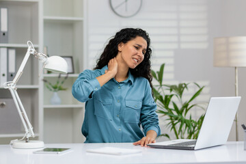 A professional woman at a desk grimaces with neck pain while working on a laptop in an office setting. - obrazy, fototapety, plakaty