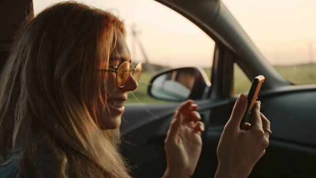 Portrait of beautiful young blonde making selfie on her phone or smartphone in front seat of the car. Pretty girl in stylish sunglasses flirting with camera while travelling. Concept of technology.