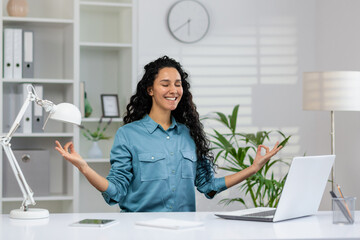 Smiling businesswoman with eyes closed practicing yoga meditation at her desk in a bright office environment to reduce stress.