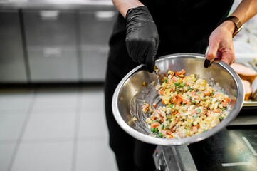 A person in black attire and gloves mixes a colorful salad in a stainless steel bowl in a modern...