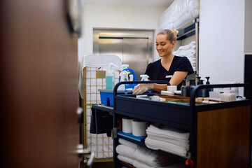 Happy housekeeper restocking room attendants' trolley with cleaning supplies in  hotel.