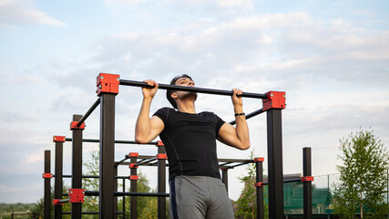 Front view of sporty man doing pull ups on horizontal bar outdoors. Young man street workout exercising at the morning time