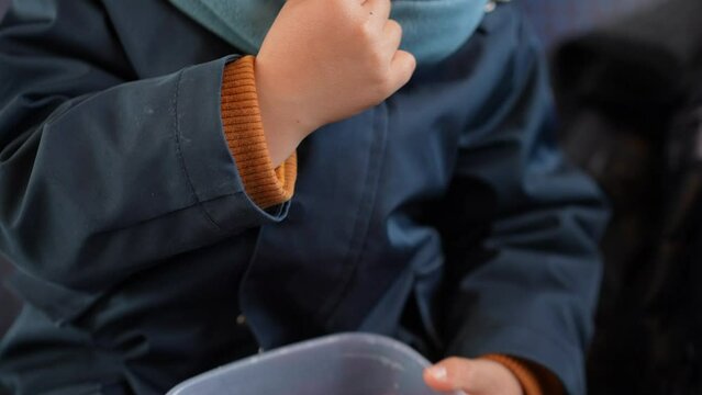 Youngster in winter attire handpicks a blueberry amidst strawberries, munching healthily from his travel container
