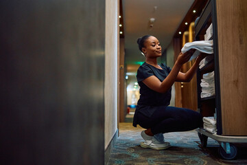 Young black maid taking clean towels from  cart while working in  hotel.