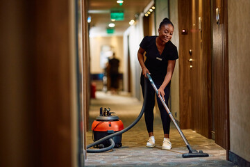 Young black maid vacuuming hallway while working in  hotel.