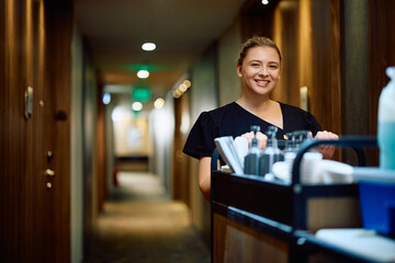 Happy housekeeper with stocked chambermaid's trolley in  hotel looking at camera.