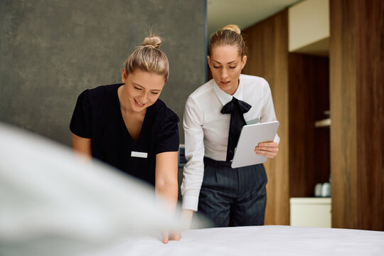 Young maid making bed with help of housekeeping supervisor in hotel room.