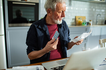 Senior man examining medication while holding documents at kitchen table with laptop and glass of water