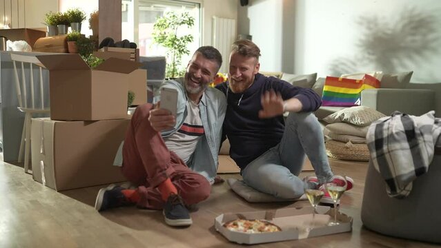 Gay couple relaxing with pizza and wine after moving into new home with boxes around