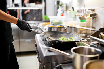 A chef in black attire sautés green vegetables in a bustling, well-equipped kitchen. Pots and pans clatter as culinary magic unfolds