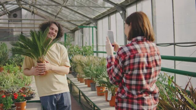 Girl photographing a man with a plant for his blog, Man and woman having fun together. Couple chose a beautiful green plant and take a pictures with it on a smartphone and are happy. Funny couple.
